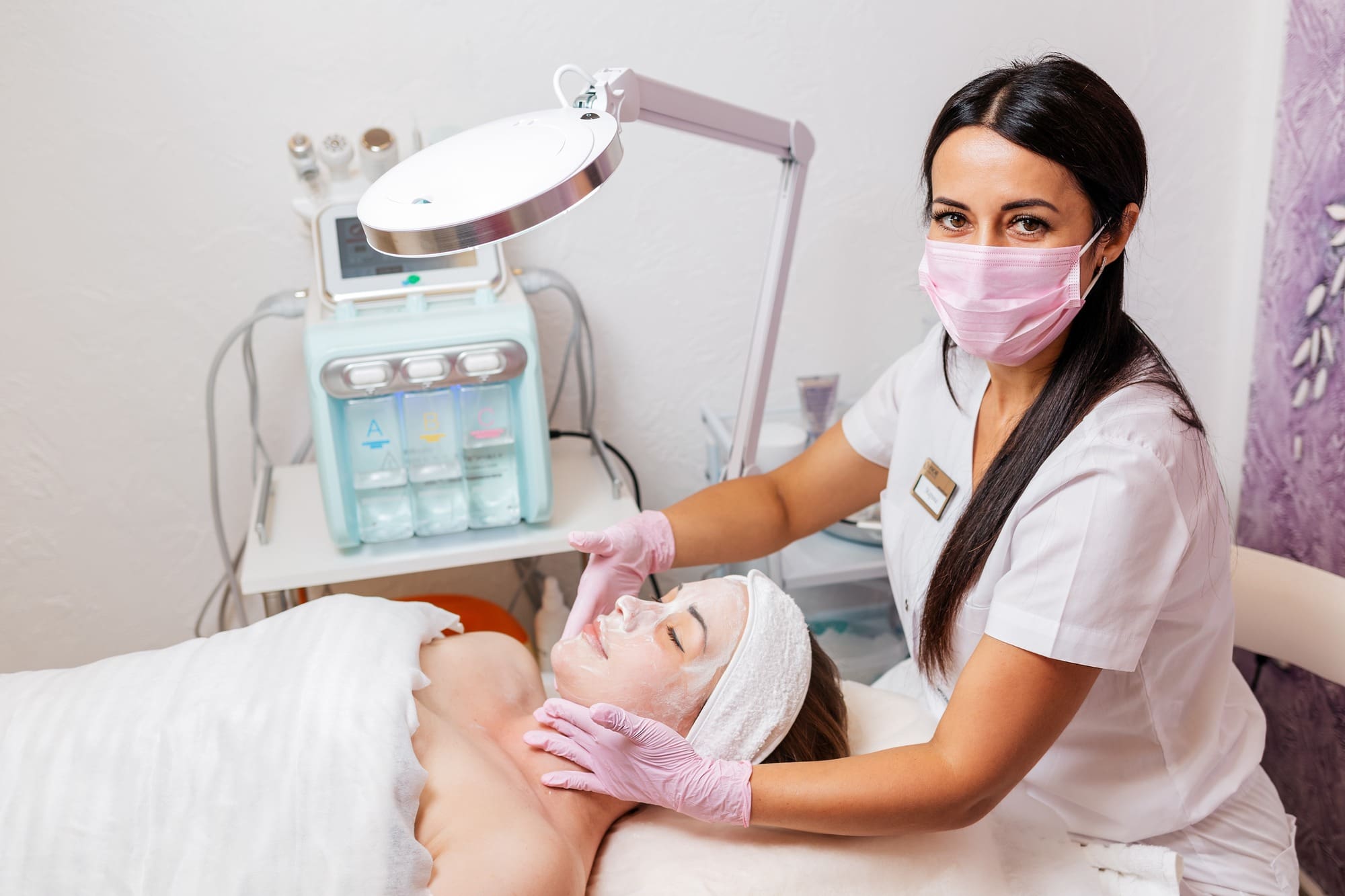 A cosmetologist in a medical mask and rubber gloves applies a cream mask to the client's face